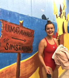 Maija Hietala punaisessa uima-asussa pyyhkeet kädessään. Taustalla on kyltti, jossa on nuoli ja siinä lukee "Uimarannalle, simstrand".