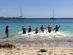 Psoriasisliiton aurinkopainotteisen sopeutumisvalmennuksen osallistujia kuvattuna takaapäin juoksemassa rannalla veteen pidellen toisiaan käsistä.