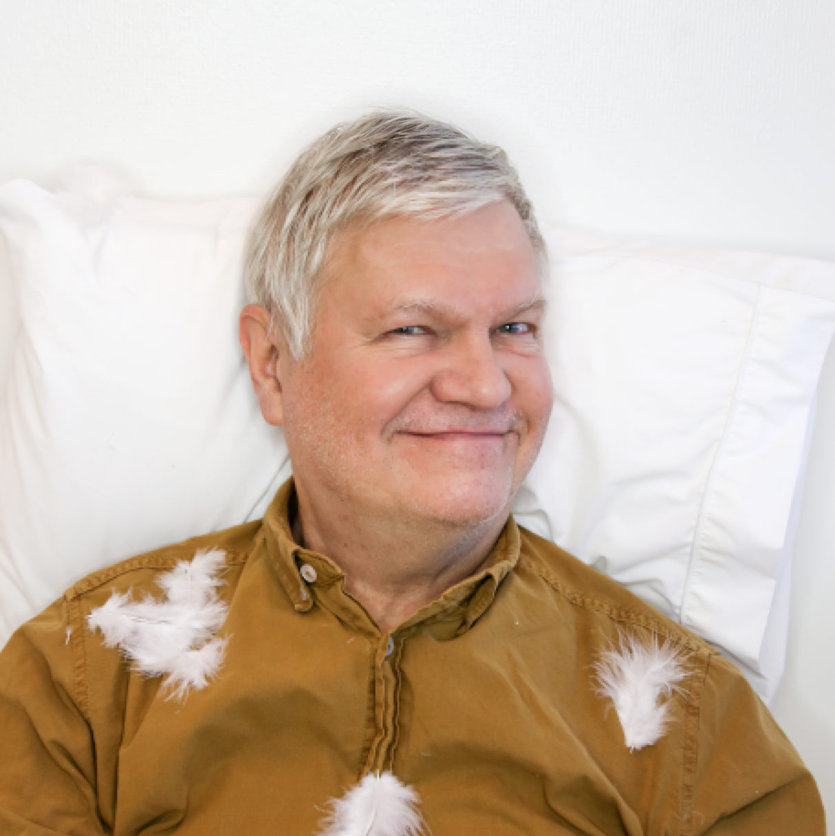 Kasvokuva Markku Partisesta, joka makaa pää tyynyllä ja hänen päällään on höyheniä.