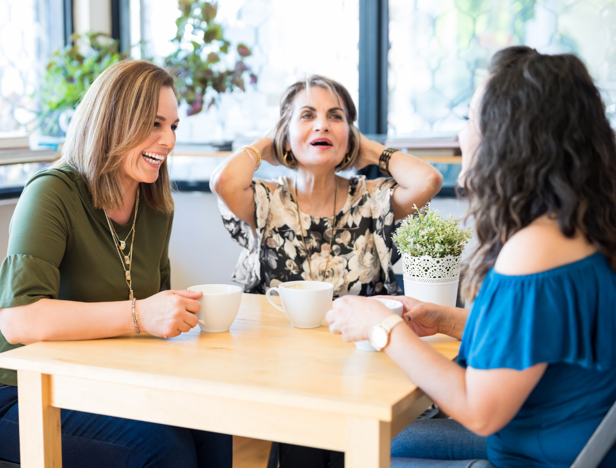 Kolme naista on kahvilla pöydän ääressä ja nauraa.