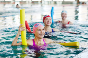 Kolme naista on uima-altaassa uimapötkylöiden kanssa ja hymyilevät.