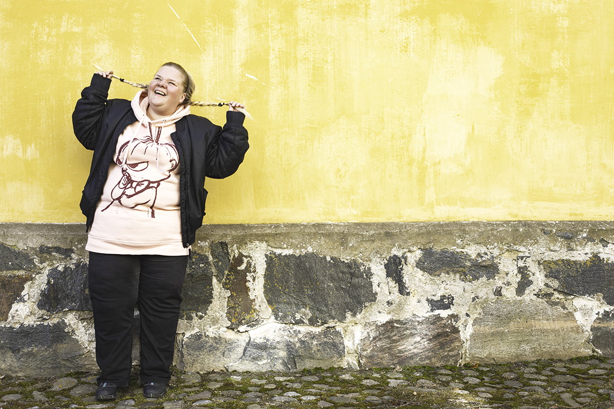 Elina Wirtanen seisoo keltaisen seinän edessä ja heiluttelee letillä olevia hiuksiaan.